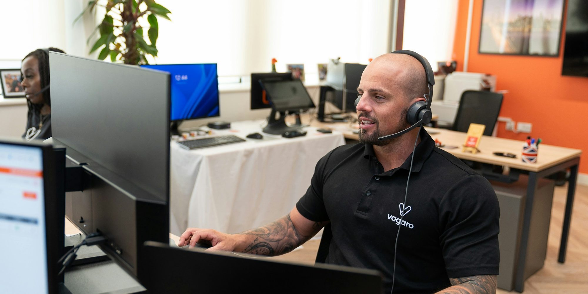 An agent wearing a telephone headset working at a desk with two monitors in an office