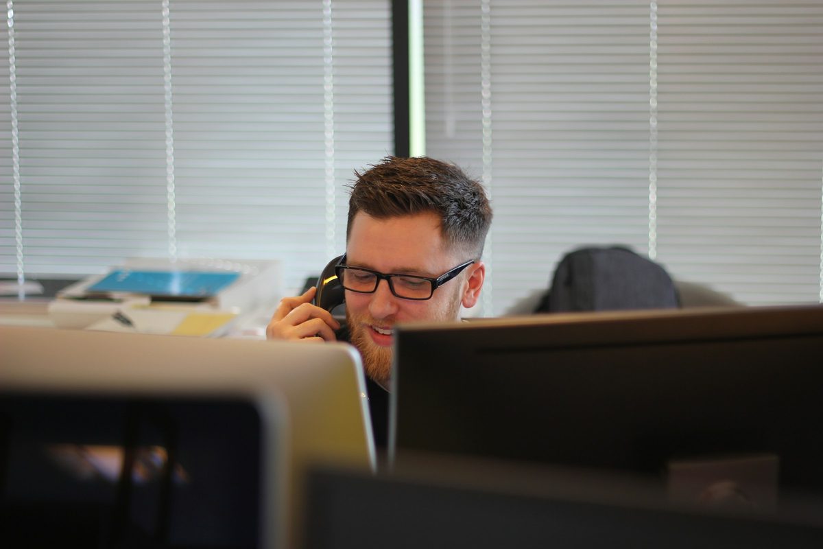A man at an office desk speaking on a telephone beside a window with venetian blinds