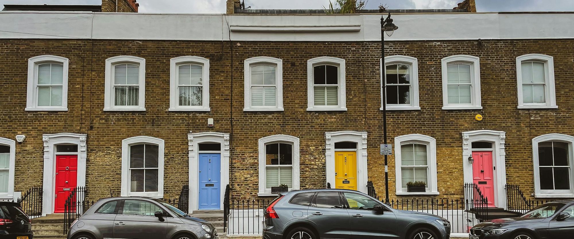 A row of British brick terraced houses with parked cars and brightly painted front doors